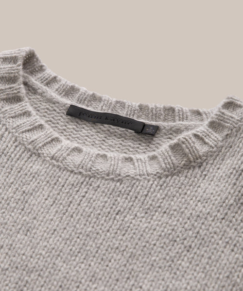 A close-up of the Cashmere Amelia Crewneck, a grey cashmere knit sweater with a ribbed crew neckline and black label inside the collar, displayed on a neutral beige background.