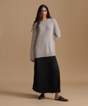 A woman modeling a light gray knit sweater and a black pleated maxi skirt against a solid beige studio background.