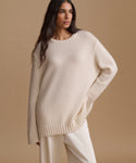 A woman wearing a cream-colored knit sweater and matching pants, photographed against a solid beige studio background.