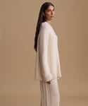 A woman stands in profile wearing a cream knit sweater and matching knit pants against a plain beige studio background.