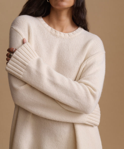 A close-up of a woman wearing a cream-colored knit sweater with a crew neck and ribbed cuffs, posed against a solid beige background.