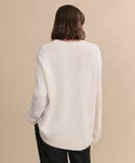 A person with shoulder-length brown hair stands with their back to the camera, wearing the oversized Cashmere Amelia Crewneck in ivory and black pants against a beige background.
