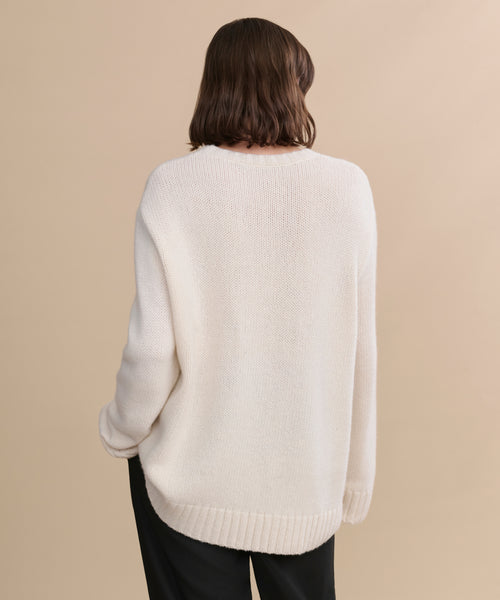 A person with shoulder-length brown hair stands with their back to the camera, wearing the oversized Cashmere Amelia Crewneck in ivory and black pants against a beige background.