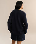 A person with long dark hair, wearing the Cashmere Amelia Crewneck in navy and dark shorts, stands facing away from the camera against a neutral beige background.