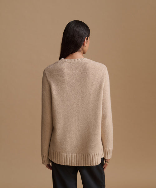 A woman viewed from the back wearing a light beige knit sweater paired with black pants, against a solid tan studio background.