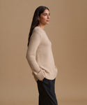 A woman in side profile wearing a light beige knit sweater and dark trousers, standing against a solid beige studio background.