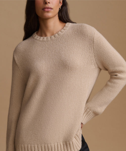 A close-up of a woman wearing a beige ribbed knit crewneck sweater against a solid warm beige background.