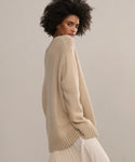 A woman with curly hair looks over her shoulder while wearing the Cashmere Amelia Crewneck and a light pleated skirt, standing against a neutral background.