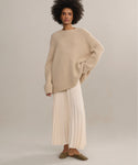 A woman with curly hair wears the Cashmere Amelia Crewneck sweater, paired with a long pleated cream skirt and brown flats, standing against a plain light backdrop.