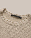 A close-up of the beige Cashmere Amelia Crewneck, a lightweight chunky knit sweater featuring a ribbed crew neckline and a black tag sewn inside the collar, displayed on a neutral background.