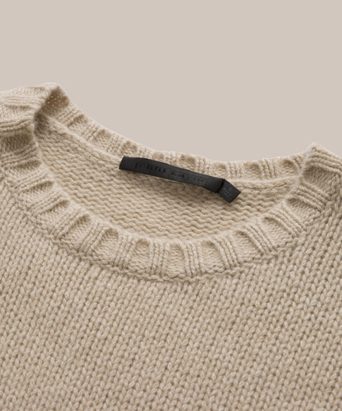 A close-up of the beige Cashmere Amelia Crewneck, a lightweight chunky knit sweater featuring a ribbed crew neckline and a black tag sewn inside the collar, displayed on a neutral background.