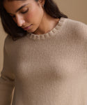 A close-up of a woman wearing a beige ribbed knit sweater, looking down, against a solid warm beige background.