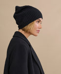 A woman with short blonde hair wears a black Cashmere Beanie and a black coat, shown in profile against a plain beige background.