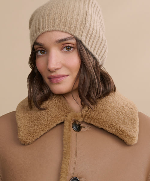 A woman with wavy brown hair wears a caramel Cashmere Beanie and a tan shearling-collared coat, looking at the camera against a neutral background.
