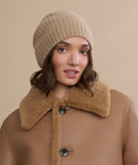 A woman in a Cashmere Beanie and a brown shearling coat with large buttons poses against a neutral beige background.