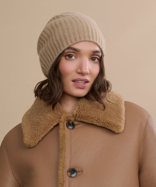 A woman in a Cashmere Beanie and a brown shearling coat with large buttons poses against a neutral beige background.