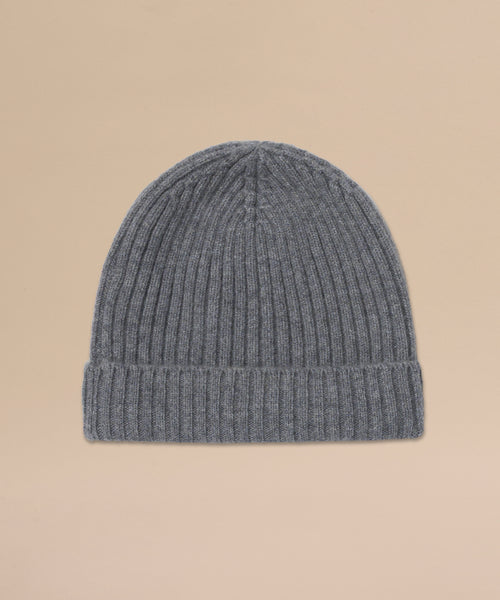 The Cashmere Beanie is a grey ribbed knit hat with a folded brim, made from lightweight cashmere and shown laid flat on a light beige background.
