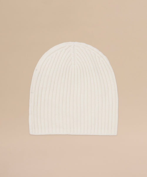 The Cashmere Beanie in ivory features a ribbed knit and lightweight cashmere blend, shown flat on a light beige background for an elegant look.