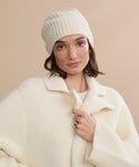 A woman wearing a Cashmere Beanie stands against a beige background in an oversized cream wool coat, looking at the camera with a neutral expression.