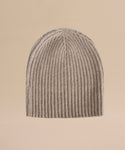 The Cashmere Beanie, featuring a beige ribbed knit design, is displayed upright against a plain light tan background.