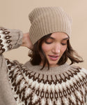 A woman wearing a Cashmere Beanie in beige and a patterned brown and white sweater looks down with one hand touching her hat against a beige background.