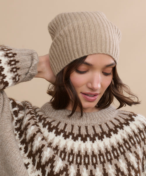 A woman wearing a Cashmere Beanie in beige and a patterned brown and white sweater looks down with one hand touching her hat against a beige background.