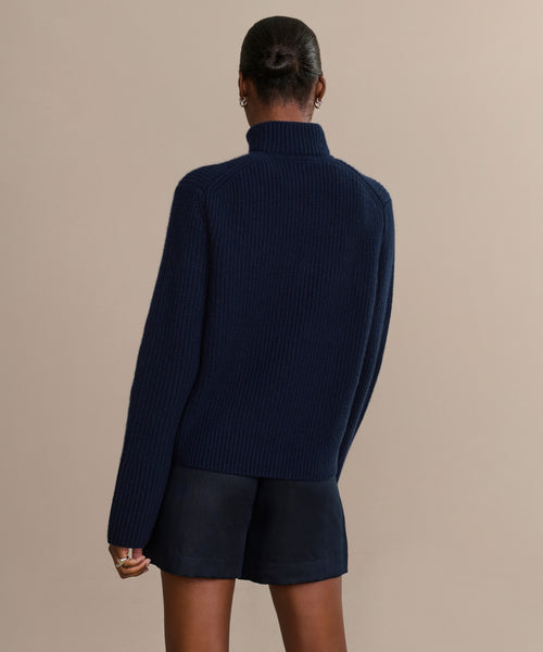 A person with their hair in a bun is shown from behind, wearing the Boyd Cashmere Sweater in dark blue and black shorts against a plain beige background.