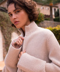 A woman with short, wavy brown hair wears the Boyd Cashmere Sweater, a beige fisherman rib zip-up style, partly unzipped to reveal a silver ring. She stands outside in sunlight with plants and buildings behind her, looking at the camera.