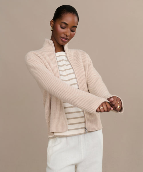 A woman wears the Boyd Cashmere Sweater in light beige over a cream and beige striped top with white pants, standing against a beige background, looking down with her arms slightly extended forward.