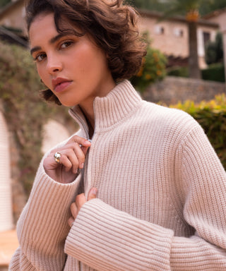 A woman with short, wavy brown hair wears the Boyd Cashmere Sweater, a beige fisherman rib zip-up style, partly unzipped to reveal a silver ring. She stands outside in sunlight with plants and buildings behind her, looking at the camera.