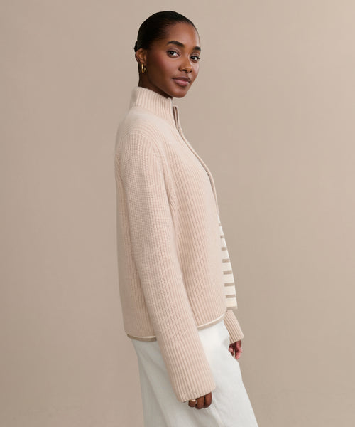 A woman with dark hair pulled back wears the Boyd Cashmere Sweater in light beige over a white top and skirt, standing sideways and looking at the camera against a plain beige background.