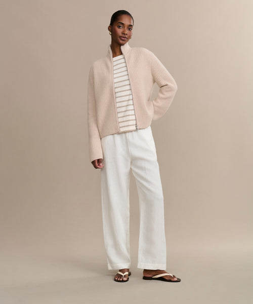 A woman stands against a beige background wearing the Boyd Cashmere Sweater in light beige over a white and beige striped top, paired with loose white pants and brown sandals. She poses with one hand in her pocket.