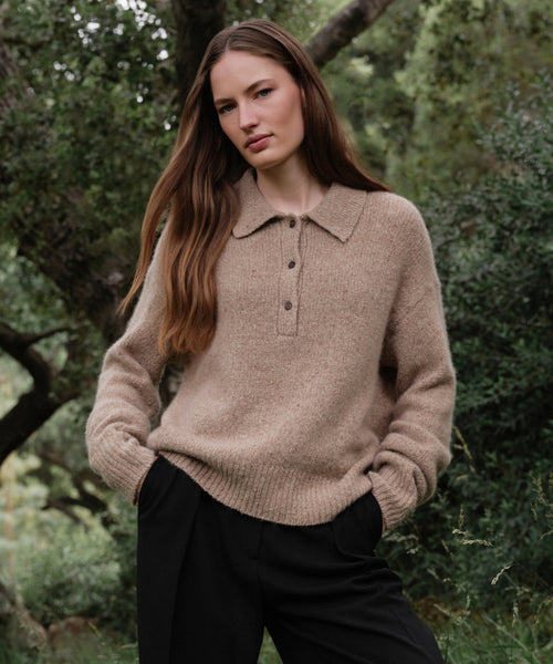 A woman with long brown hair stands outdoors among green trees, wearing the Cashmere Burke Polo and black pants, her hands in her pockets and a calm expression on her face.