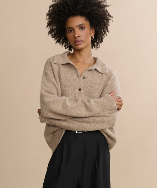 A person with curly hair wears the Cashmere Burke Polo, styled oversized with large sleeves and black pants, standing with arms crossed against a plain beige background.
