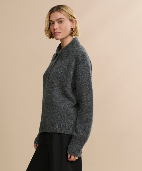 A woman with short blonde hair wears an oversized, dark gray Cashmere Burke Polo sweater with a collar and long sleeves, paired with black pants. She stands against a plain beige background, looking slightly to the side.