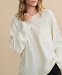 A woman with wavy blonde hair poses against a beige background, wearing the off-white Cashmere Chandler Long-Sleeve Tee featuring a chest pocket. Her face is partly out of frame as she lightly touches her collar with one hand.