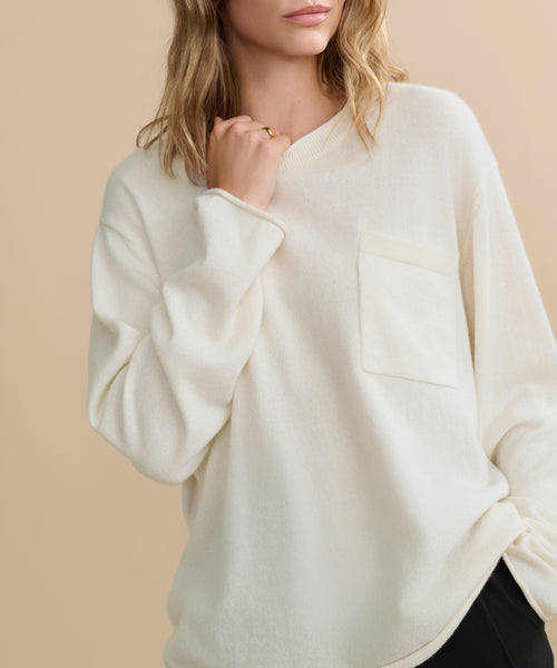 A woman with wavy blonde hair poses against a beige background, wearing the off-white Cashmere Chandler Long-Sleeve Tee featuring a chest pocket. Her face is partly out of frame as she lightly touches her collar with one hand.