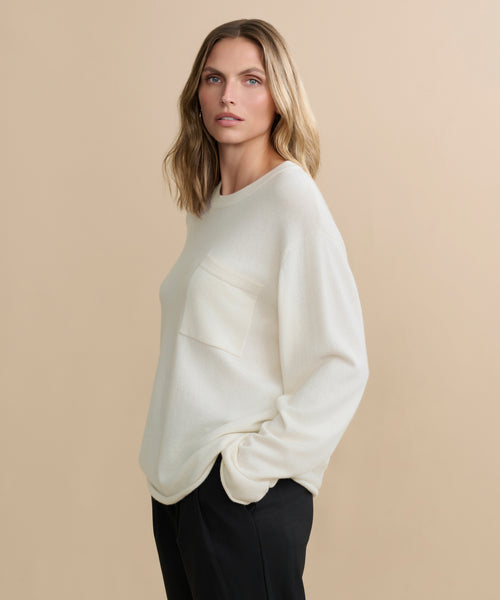 A woman with shoulder-length blonde hair wears the Cashmere Chandler Long-Sleeve Tee featuring a chest pocket, paired with black pants. She stands with one hand in her pocket against a plain beige background.