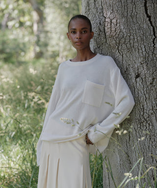 A person with closely-cropped hair wears the Cashmere Chandler Long-Sleeve Tee with a pocket and a light skirt while leaning against a tree in a grassy, sunlit outdoor setting.