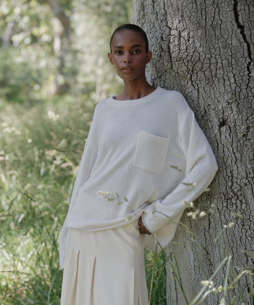 A person with closely-cropped hair wears the Cashmere Chandler Long-Sleeve Tee with a pocket and a light skirt while leaning against a tree in a grassy, sunlit outdoor setting.