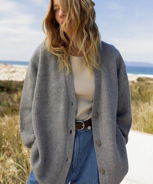 A woman wearing an oversized gray ribbed cardigan over a cream top and blue jeans, standing outdoors near a beach with grass and sky in the background.