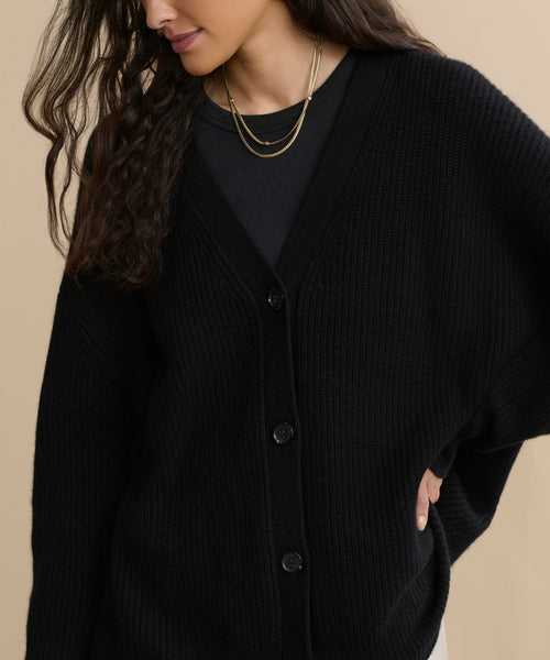 A woman models the Cashmere Cocoon Cardigan in black over a matching top, styled with layered gold necklaces. She poses with one hand on her hip, her long wavy dark hair contrasting against a beige background.