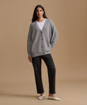 A woman stands against a plain beige studio background wearing a light gray knit cardigan over a white top, black pants, and off-white flats.