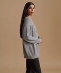 A woman modeling an oversized light gray ribbed knit sweater with black pants, standing against a solid beige studio background.