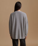 A woman viewed from the back wearing an oversized light gray ribbed knit sweater and black pants, standing against a plain warm beige studio background.