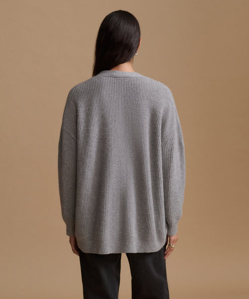 A woman viewed from the back wearing an oversized light gray ribbed knit sweater and black pants, standing against a plain warm beige studio background.