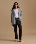 A woman stands against a plain beige studio background wearing a light gray cardigan over a white crew-neck top, black straight-leg jeans, and off-white flats.