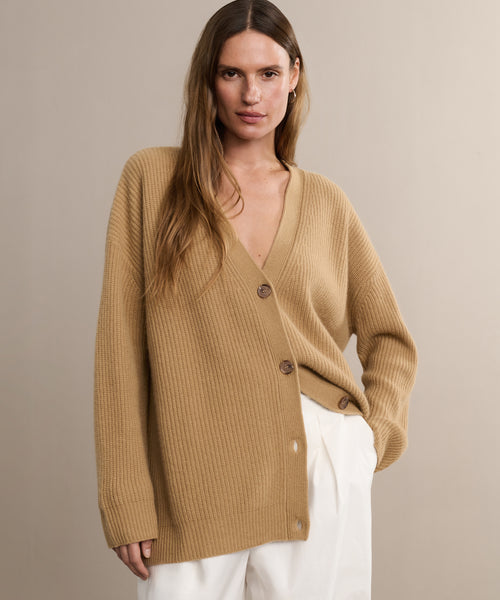 A woman with long brown hair wears the Cashmere Cocoon Cardigan, a loose, slouchy style with three buttons, paired with white pants, standing against a plain beige background.