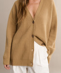 A person wears the Cashmere Cocoon Cardigan—a slouchy, tan ribbed knit with brown buttons—partially unbuttoned over high-waisted, wide-leg white pants, with one hand tucked in their pocket.