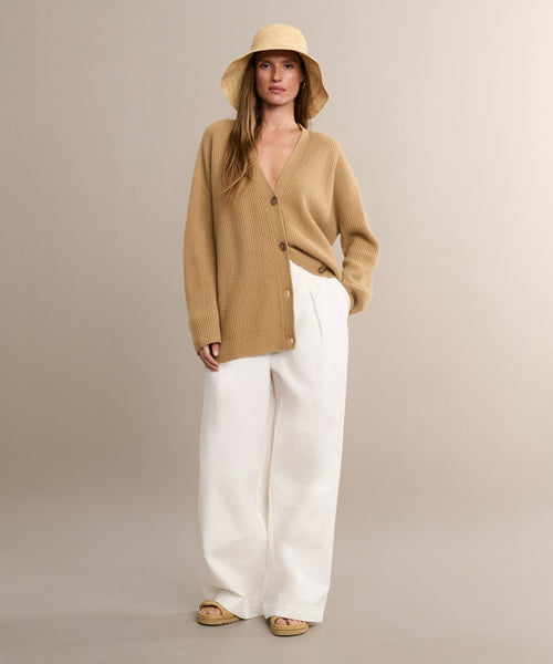A woman wears a tan wide-brim hat, the Cashmere Cocoon Cardigan, white wide-leg pants, and beige sandals while standing against a neutral background.